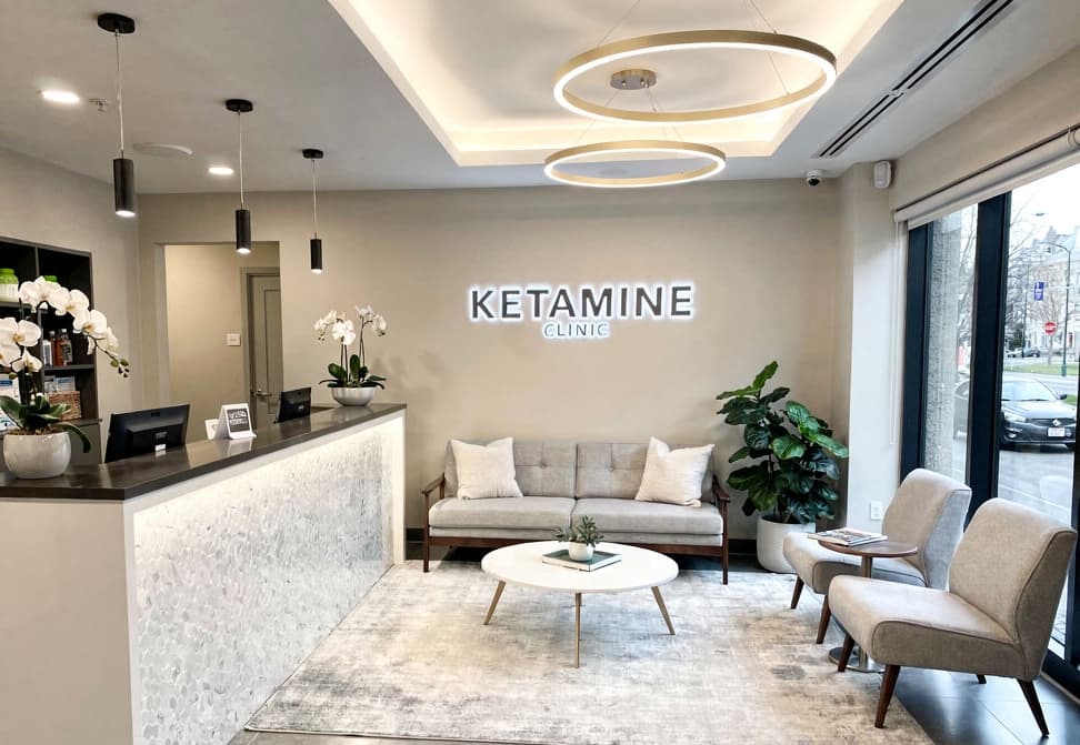 A modern waiting area in a ketamine clinic with a sofa, two chairs, coffee table, potted plants, and reception counter—perfect for Ketamine Clinic Digital Marketing photos. A "KETAMINE CLINIC" sign adorns the wall and large windows fill the space with natural light. - Ketamine