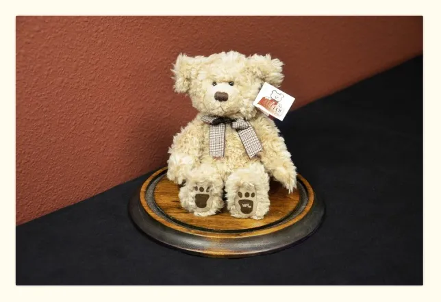 A plush teddy bear with a checkered bow tie sits on a round wooden base against a red wall, offering comfort akin to that sought by ketamine therapy patients. The bear's paws feature paw print designs, and a small tag is attached to its ear. The base rests on a dark surface. - Ketamine