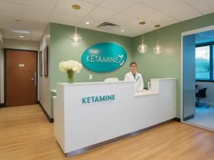 A receptionist in a white coat stands behind a white desk, welcoming ketamine therapy patients. The clinic, with its green walls and 
