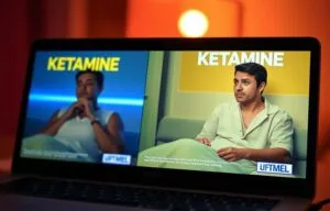 A laptop screen displays two side-by-side images of a man with “KETAMINE” in large text above each, illustrating Ketamine therapy. The man is in different settings with unique shirts. Subtitles and a “UFTMEL” logo appear at the bottom. - Ketamine