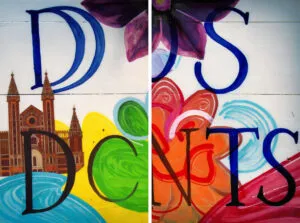 A mural features large blue letters, colorful abstract shapes, a purple flower, and an illustration of a cathedral. Inspired by artificial intelligence, the design is divided in the middle by a white line, creating a split effect. - Ketamine