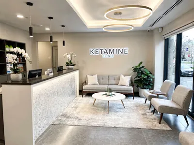 A modern waiting room with a reception desk, neutral-colored chairs, a couch, coffee table, decorative plants, and a "Ketamine Clinic" sign. Large windows let in natural light—an inviting space perfect for Ketamine Clinic digital marketing visuals. - Ketamine